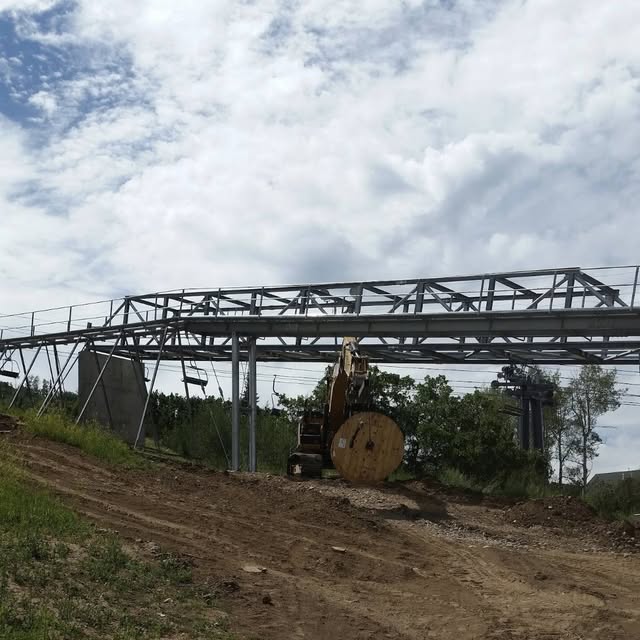 A steel chairlift line structure under construction with a large cable spool on the mountainside