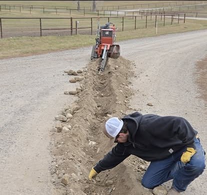 A worker at an open trench with a ride-on trencher running underground service