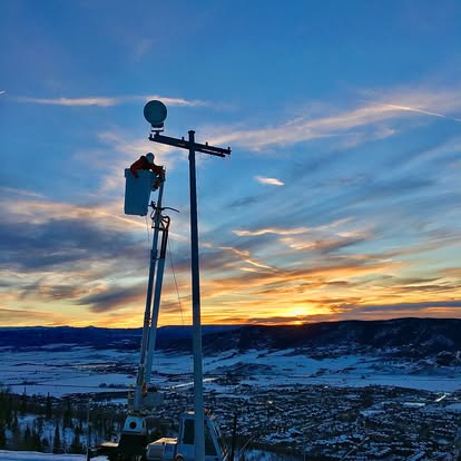 A Current Electric worker in a bucket-truck lift working at the top of a pole above the snowy Steamboat valley at sunset
